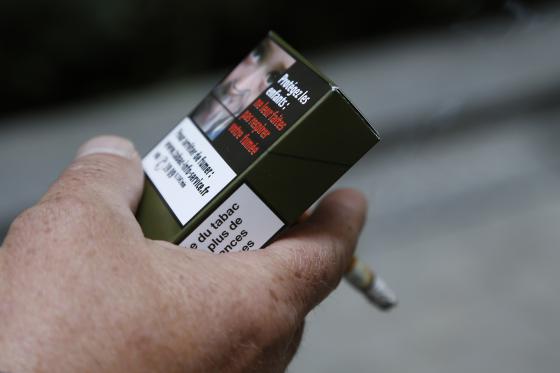 A man smokes a cigarette on September 25, 2014 in Paris, holding a sample of a "plain cigarette packaging" cigarette box proposed by the "Alliance contre le tabac" (alliance against tobacco) association. France on September 25 said it would introduce plain cigarette packaging and ban electronic cigarettes in certain public places, in a bid to reduce high smoking rates among the under-16s. Following a successful similar campaign in Australia, Health Minister Marisol Touraine said cigarette packets would be "the same shape, same size, same colour, same typeset" to make smoking less attractive to young smokers. AFP PHOTO THOMAS SAMSON (Photo credit should read THOMAS SAMSON/AFP/Getty Images)