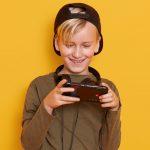 Horizontal shot of little boy wearing black cap and green hoody, posing with cell phone in hands, handsome child isolated over yellow studio background, fashionable kid playing online games.